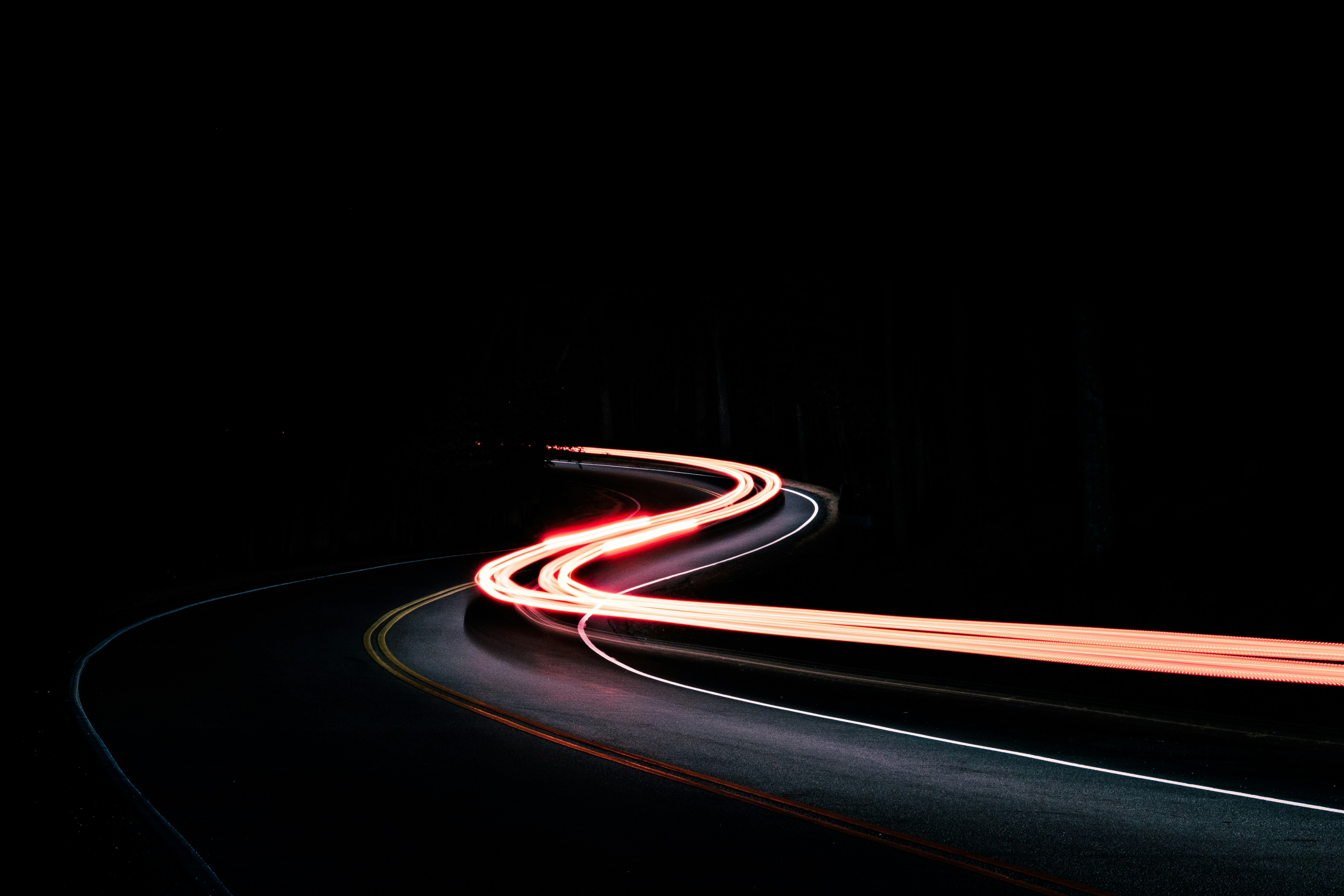 Long exposure photo of a winding road at night with red and white light trails from a fast-moving vehicle.